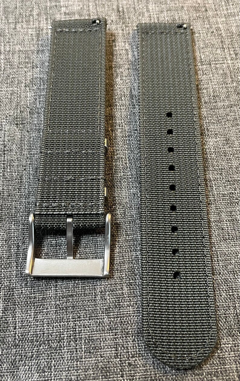 K&ouml;nnte beinhalten: Zwei dunkelgraue Nylon-Uhrenarmb&auml;nder mit silbernen Schlie&szlig;en. Die Armb&auml;nder sind aus gewebtem Material und haben eine strukturierte Oberfl&auml;che.