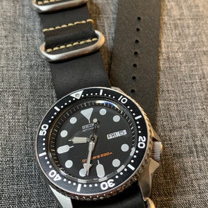 以下が含まれることがあります： 黒と銀のセイコー自動巻き腕時計、黒い革のストラップ付き。時計の文字盤は黒地に白の目盛り、日付表示窓には「FRI 5」と表示されています。この時計は200メートル防水です。