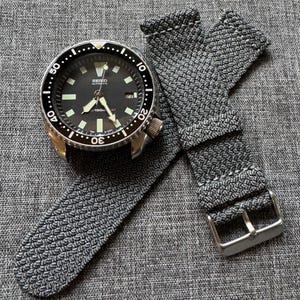 Peut inclure: Une montre automatique Seiko en acier inoxydable avec un cadran noir et un bracelet en nylon tissé gris. La montre dispose d'une lunette rotative et d'aiguilles et d'index luminescents. Le bracelet a une boucle argentée.