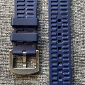 Puede incluir: Correa de reloj azul marino de dos piezas con hebilla plateada. La correa est&aacute; hecha de un material flexible con un dise&ntilde;o texturizado y acanalado. Dise&ntilde;ada para un reloj, se muestra sobre una superficie gris.