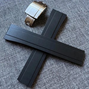 Puede incluir: Un cierre de reloj de acero inoxidable y dos correas de reloj de goma negra se muestran sobre una superficie gris texturizada. El cierre del reloj es rectangular con un acabado de metal cepillado. Las correas del reloj son planas y rectangulares.