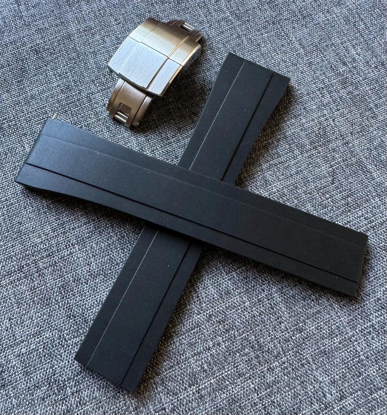 Puede incluir: Un cierre de reloj de acero inoxidable y dos correas de reloj de goma negra se muestran sobre una superficie gris texturizada. El cierre del reloj es rectangular con un acabado de metal cepillado. Las correas del reloj son planas y rectangulares.