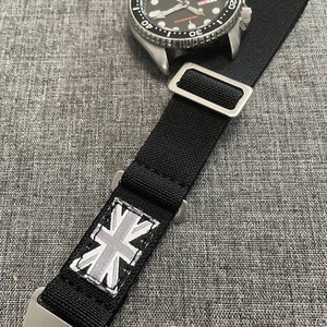 May include: A black watch strap with a white Union Jack flag design. The strap is made of nylon and has a silver buckle. The strap is attached to a silver watch with a black face and a silver bezel.