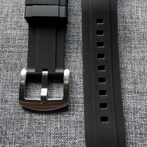 May include: Black watch band with a stainless steel buckle. The band is made of a flexible material and features a series of rectangular holes. The band is designed to attach to a watch case.
