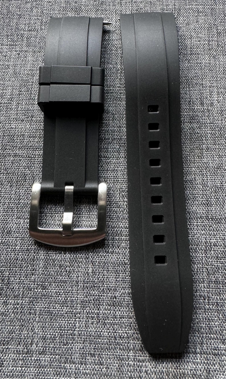 May include: Black watch band with a stainless steel buckle. The band is made of a flexible material and features a series of rectangular holes. The band is designed to attach to a watch case.