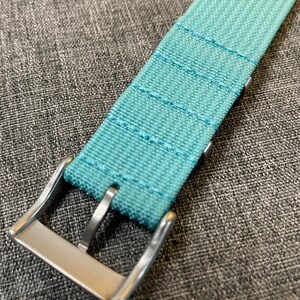 May include: A light blue watch strap with a silver buckle. The strap is made of a woven fabric with a subtle texture.