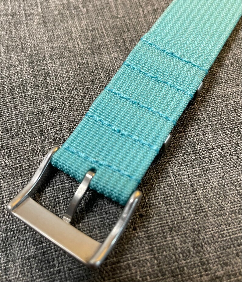 May include: A light blue watch strap with a silver buckle. The strap is made of a woven fabric with a subtle texture.