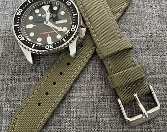 May include: A black Seiko Diver's 200m watch with a black bezel and a green canvas watch strap. The watch face displays the date and time. The strap has a silver buckle and white stitching.