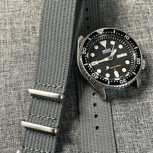 May include: A black Seiko automatic diver's watch with a gray nylon watch strap. The watch face has white markings and a date window that reads "TUE 4".