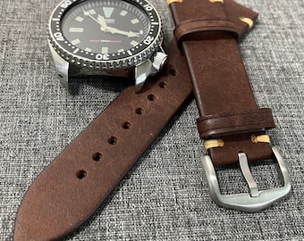 Può includere: Un cinturino da orologio in pelle marrone con cuciture bianche e una fibbia argentata. Il cinturino è mostrato con un quadrante dell'orologio nero con una ghiera argentata.
