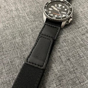 May include: A black and silver wristwatch with a black band featuring a white and blue American flag design. The watch face is black with white and orange accents. The watch is on a gray textured surface.