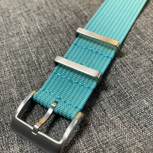 May include: A light blue watch strap with a silver buckle. The strap is made of a woven fabric.