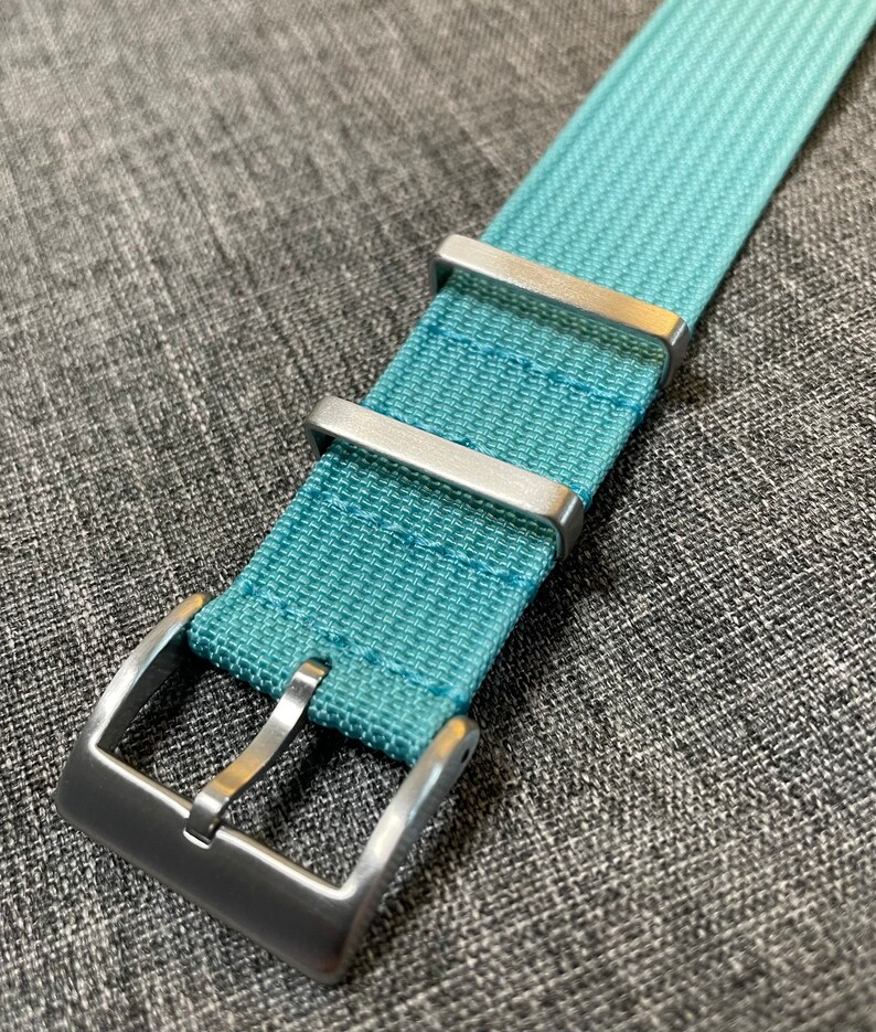 May include: A light blue watch strap with a silver buckle. The strap is made of a woven fabric.