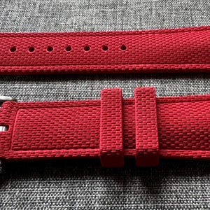 Puede incluir: Una correa de reloj roja con una superficie texturizada y una hebilla plateada. La correa tiene m&uacute;ltiples agujeros para ajustar y dos presillas para asegurar el exceso de longitud. La correa del reloj se muestra sobre un fondo gris.