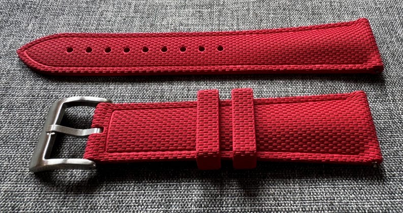 Puede incluir: Una correa de reloj roja con una superficie texturizada y una hebilla plateada. La correa tiene m&uacute;ltiples agujeros para ajustar y dos presillas para asegurar el exceso de longitud. La correa del reloj se muestra sobre un fondo gris.