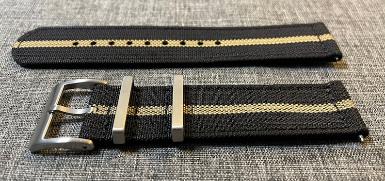 May include: A black watch strap with two thin beige stripes and a silver buckle.