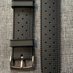 Puede incluir: Una correa de reloj de caucho negro con un patr&oacute;n texturizado y agujeros para la ventilaci&oacute;n. La correa tiene una hebilla plateada.