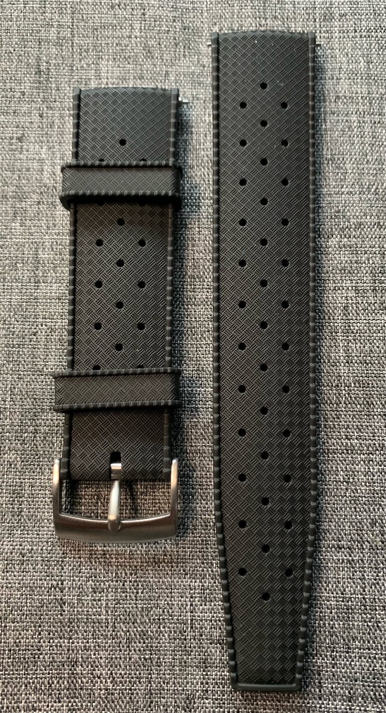 Puede incluir: Una correa de reloj de caucho negro con un patr&oacute;n texturizado y agujeros para la ventilaci&oacute;n. La correa tiene una hebilla plateada.