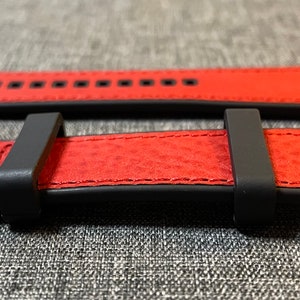 Op de afbeelding: Een rood leren horlogebandje met zwarte stiksels en een zilveren gesp. Het bandje wordt in twee posities getoond, een met de gesp gesloten en een met de gesp open.