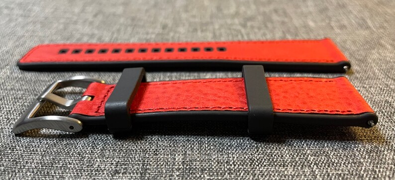Op de afbeelding: Een rood leren horlogebandje met zwarte stiksels en een zilveren gesp. Het bandje wordt in twee posities getoond, een met de gesp gesloten en een met de gesp open.