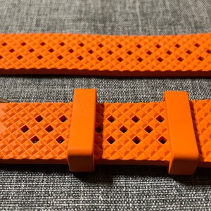 Puede incluir: Correa de reloj naranja con un patr&oacute;n de diamante texturizado y una hebilla plateada.