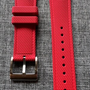 Puede incluir: Un par de correas de reloj de color rojo brillante con un patr&oacute;n texturizado. Una correa tiene una hebilla de color plateado y la otra tiene varios agujeros para el ajuste. Las correas se muestran sobre una superficie gris texturizada.
