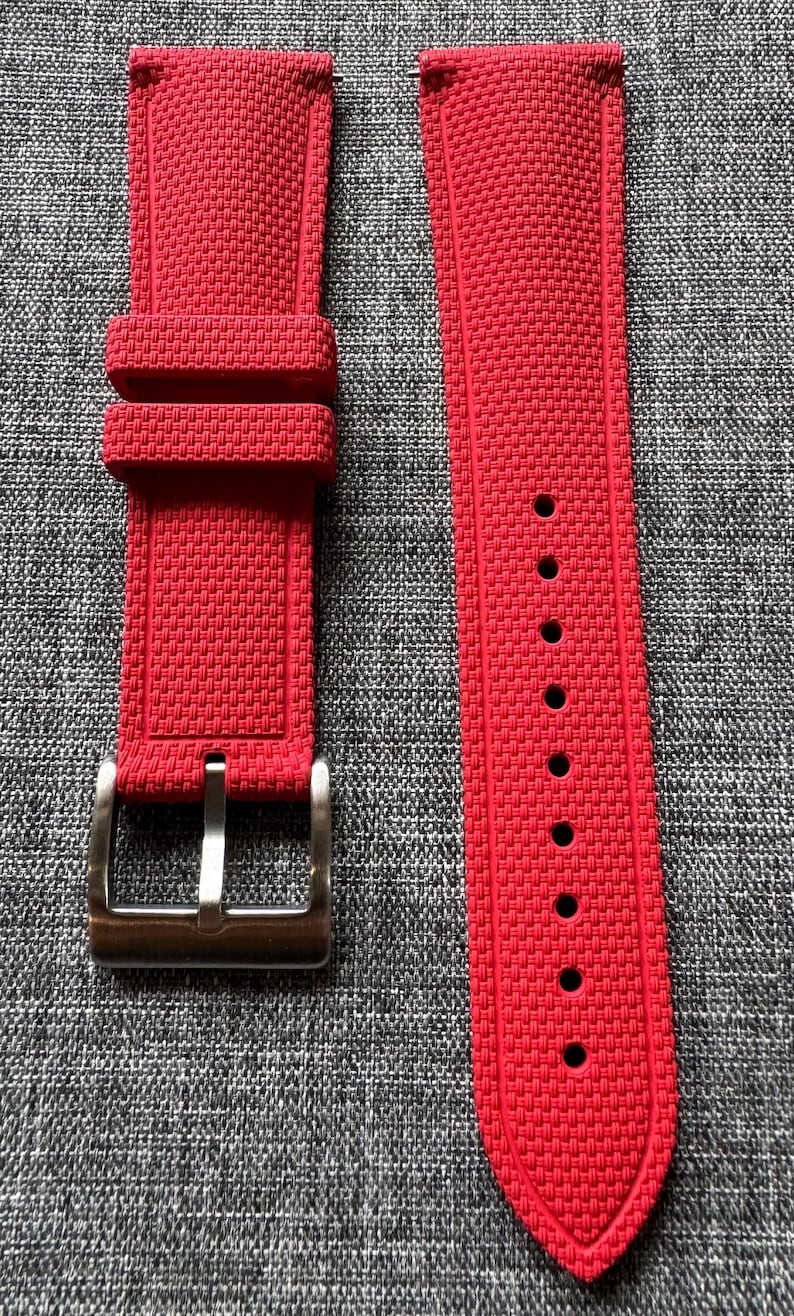 Puede incluir: Un par de correas de reloj de color rojo brillante con un patr&oacute;n texturizado. Una correa tiene una hebilla de color plateado y la otra tiene varios agujeros para el ajuste. Las correas se muestran sobre una superficie gris texturizada.