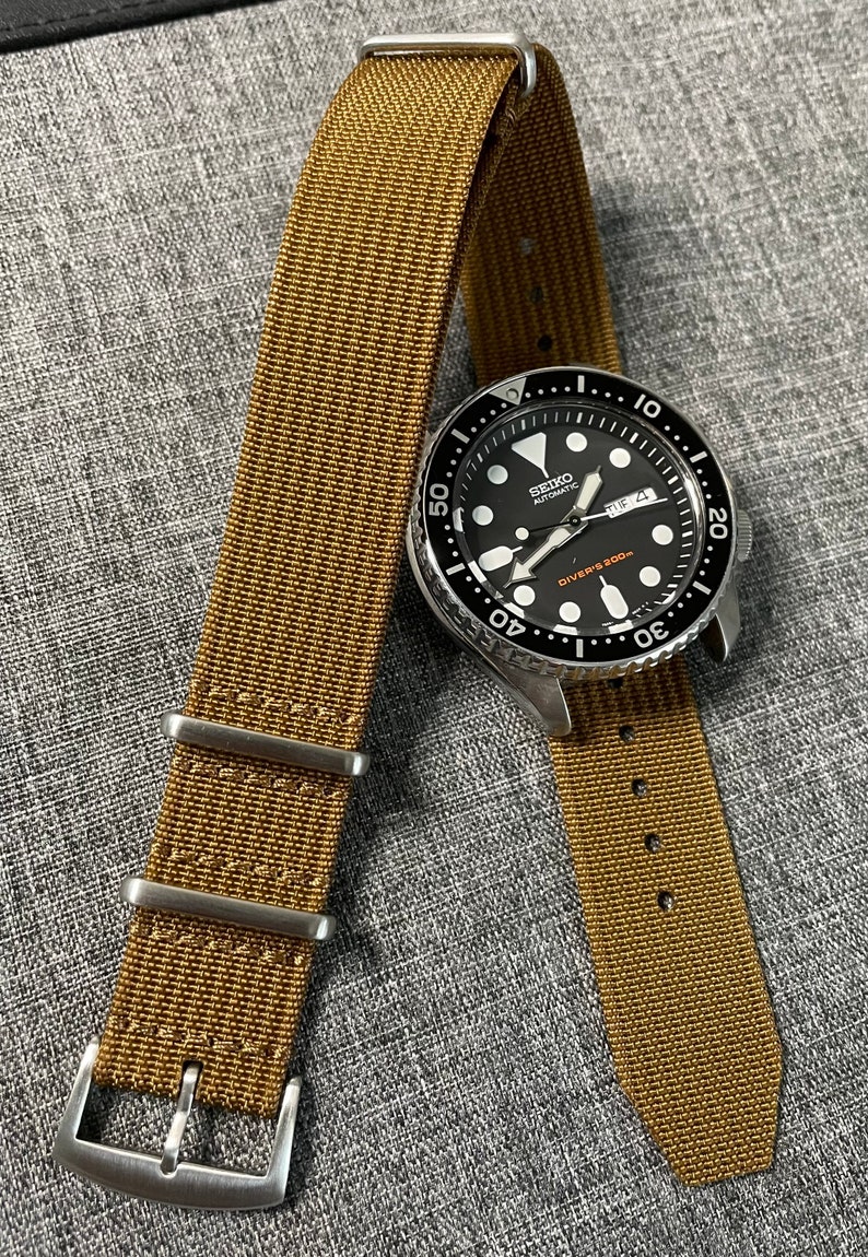 K&ouml;nnte beinhalten: Eine silberne Seiko Automatikuhr mit schwarzem Zifferblatt und einem braunen Nylon-Armband. Das Zifferblatt der Uhr hat wei&szlig;e Markierungen und den Text "SEIKO AUTOMATIC DIVERS 200m".