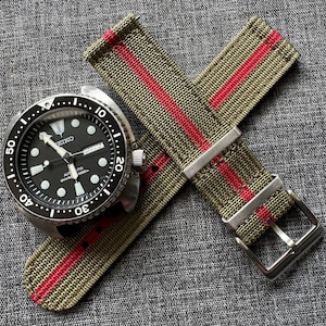 May include: A green and red striped nylon watch strap with a silver buckle. The strap is shown on top of a black watch with a white face and silver hands.