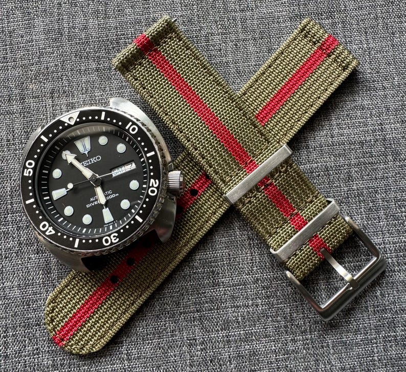 May include: A green and red striped nylon watch strap with a silver buckle. The strap is shown on top of a black watch with a white face and silver hands.