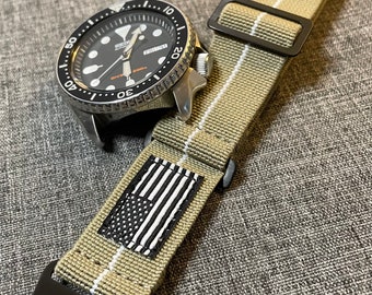 May include: A black and white American flag patch on a khaki watch strap with white stripes. The strap has a black buckle with the letters "U.S." on it.