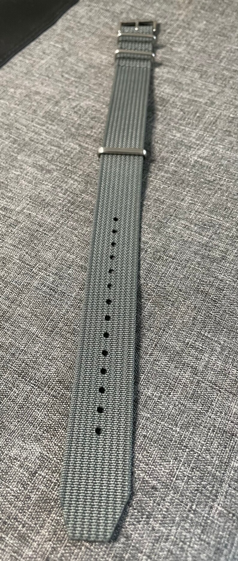 May include: A gray woven watch strap with silver hardware and multiple holes for adjusting the size.