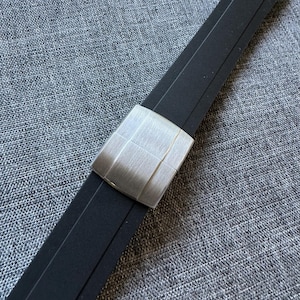 Puede incluir: Una correa de reloj negra con un cierre de metal plateado cepillado. El cierre es rectangular con un dise&ntilde;o de rejilla. La correa est&aacute; hecha de un material flexible y est&aacute; sobre un fondo gris texturizado.