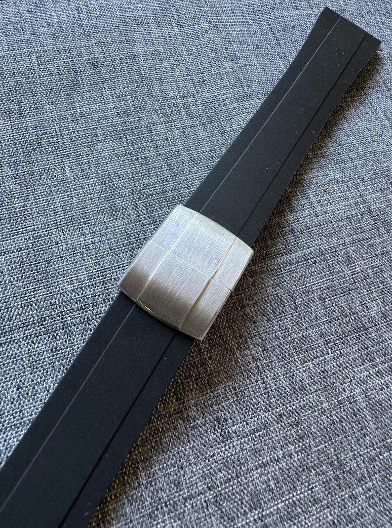 Puede incluir: Una correa de reloj negra con un cierre de metal plateado cepillado. El cierre es rectangular con un dise&ntilde;o de rejilla. La correa est&aacute; hecha de un material flexible y est&aacute; sobre un fondo gris texturizado.