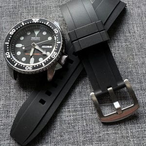 May include: A black Seiko automatic dive watch with a black rubber strap. The watch has a black dial with white markers and hands. The strap has a silver buckle. The watch is displayed on a gray textured surface.