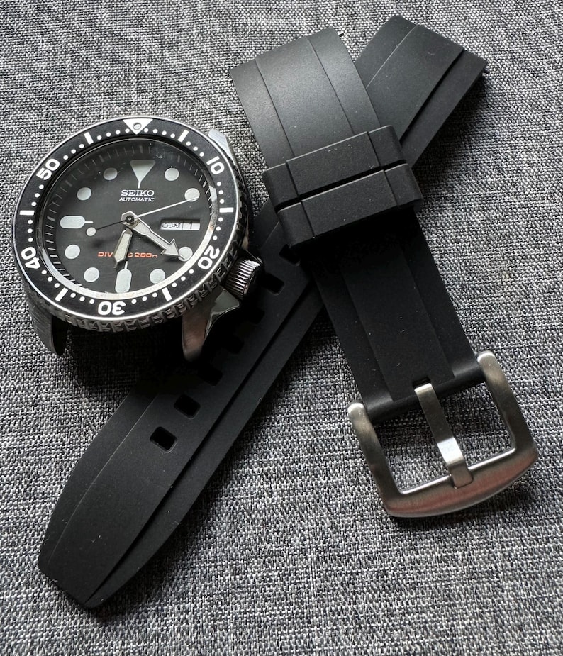 May include: A black Seiko automatic dive watch with a black rubber strap. The watch has a black dial with white markers and hands. The strap has a silver buckle. The watch is displayed on a gray textured surface.