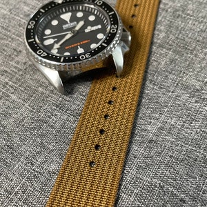 K&ouml;nnte beinhalten: Eine silberne Uhr mit schwarzem Zifferblatt und einem braunen Nylonarmband. Das Zifferblatt der Uhr hat wei&szlig;e Markierungen und den Text "SEIKO AUTOMATIC DIVERS 200m".