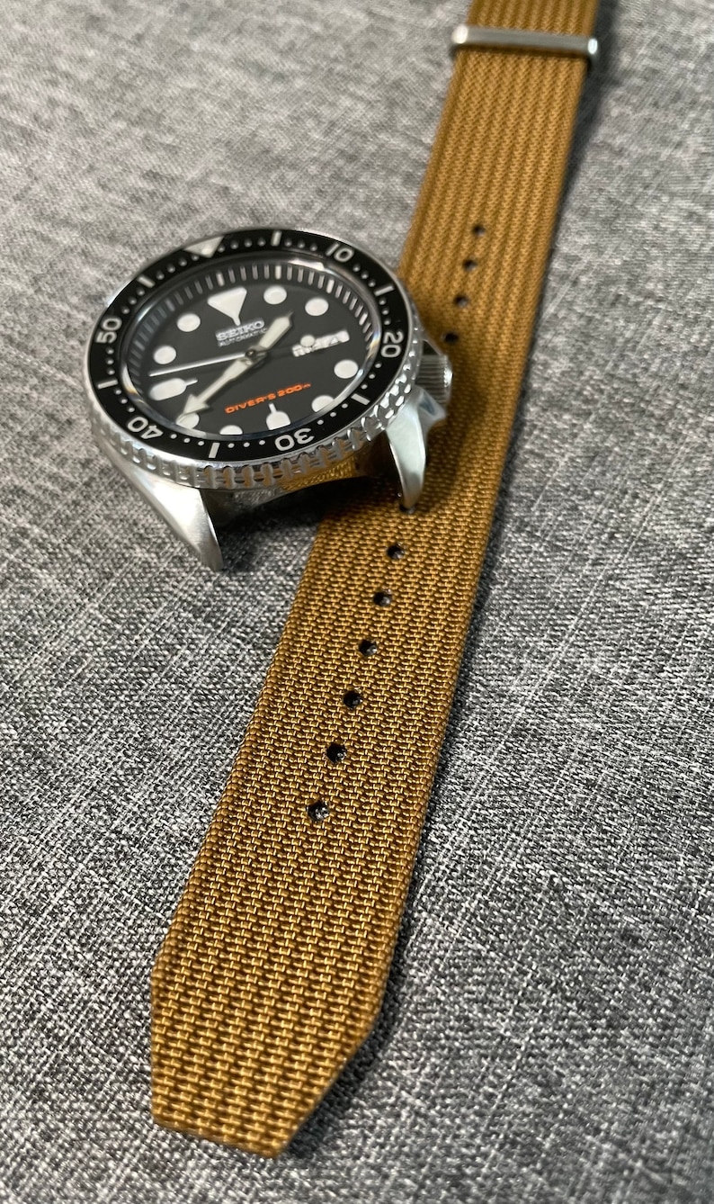 K&ouml;nnte beinhalten: Eine silberne Uhr mit schwarzem Zifferblatt und einem braunen Nylonarmband. Das Zifferblatt der Uhr hat wei&szlig;e Markierungen und den Text "SEIKO AUTOMATIC DIVERS 200m".