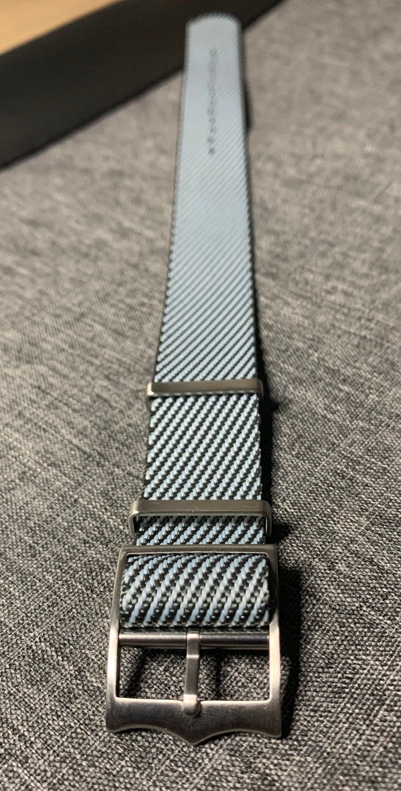 May include: A black and white striped watch strap with a silver buckle. The strap is made of a woven material and has a textured surface.