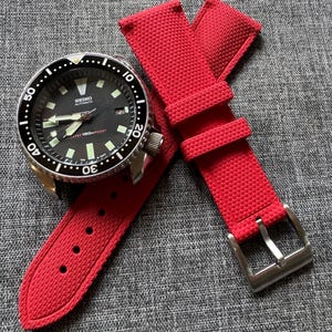 Puede incluir: Un reloj autom&aacute;tico Seiko negro con bisel plateado y correa roja. La esfera del reloj tiene marcadores y agujas blancas. La correa roja tiene una superficie texturizada y una hebilla plateada.