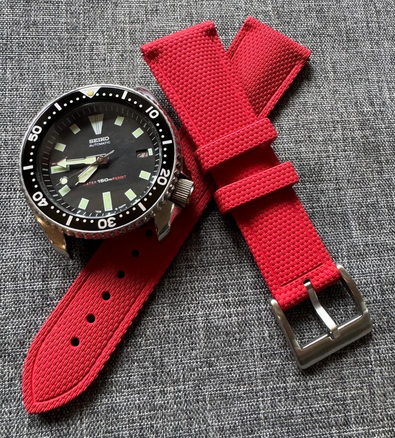 Puede incluir: Un reloj autom&aacute;tico Seiko negro con bisel plateado y correa roja. La esfera del reloj tiene marcadores y agujas blancas. La correa roja tiene una superficie texturizada y una hebilla plateada.