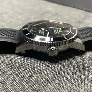 May include: A black and silver wristwatch with a black rubber strap. The watch has a round face with a black dial and silver hands. The watch is on a grey surface.