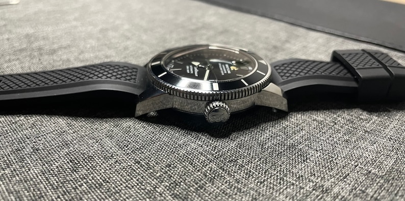 May include: A black and silver wristwatch with a black rubber strap. The watch has a round face with a black dial and silver hands. The watch is on a grey surface.