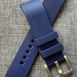 Puede incluir: Una correa de reloj azul marino con una hebilla plateada. La correa est&aacute; hecha de un material flexible y presenta cortes cuadrados cerca del extremo. La hebilla es rectangular con un acabado de metal cepillado.