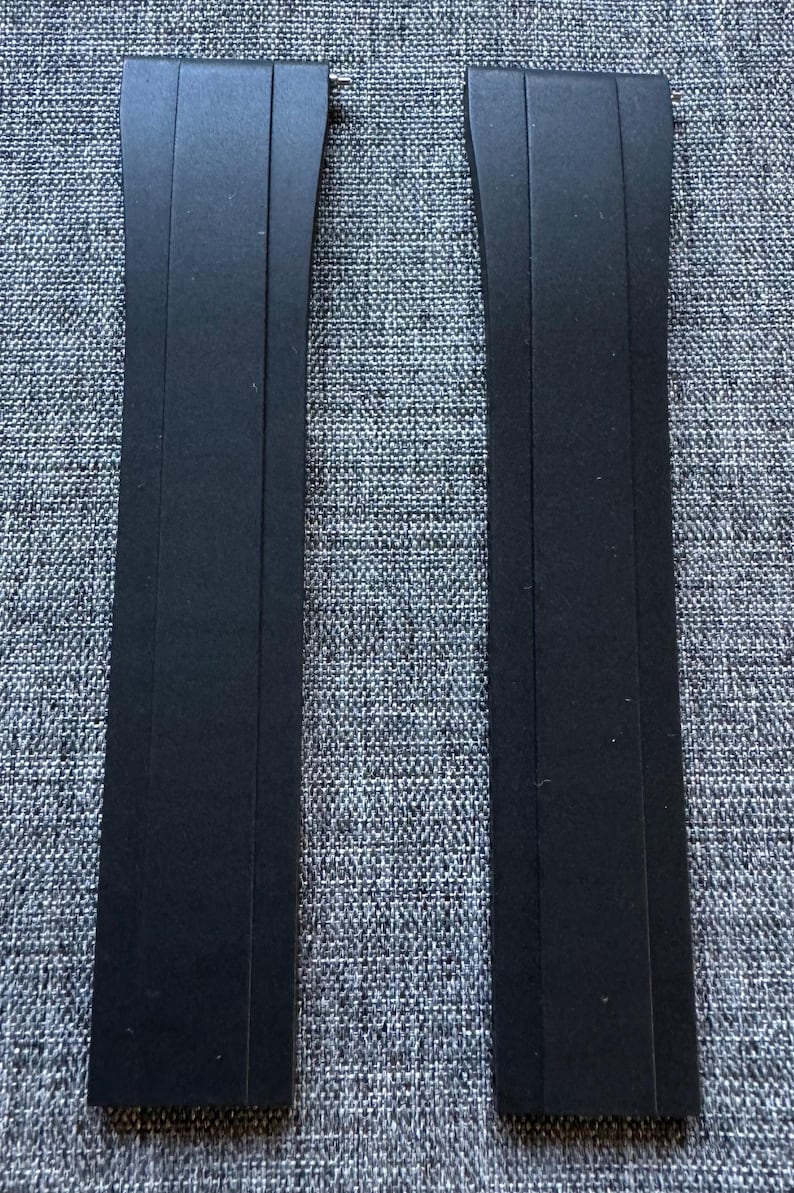 Puede incluir: Dos correas de reloj negras se muestran sobre un fondo gris texturizado. Cada correa es rectangular con un extremo curvo y un pasador de metal para la fijaci&oacute;n. Las correas est&aacute;n hechas de un material mate.