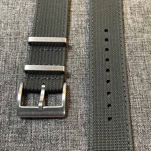 K&ouml;nnte beinhalten: Zwei schwarze Nylon-Uhrenarmb&auml;nder mit silbernen Schlie&szlig;en. Die Armb&auml;nder sind flach und haben eine gewebte Textur. Die Schlie&szlig;en haben eine geb&uuml;rstete Oberfl&auml;che.