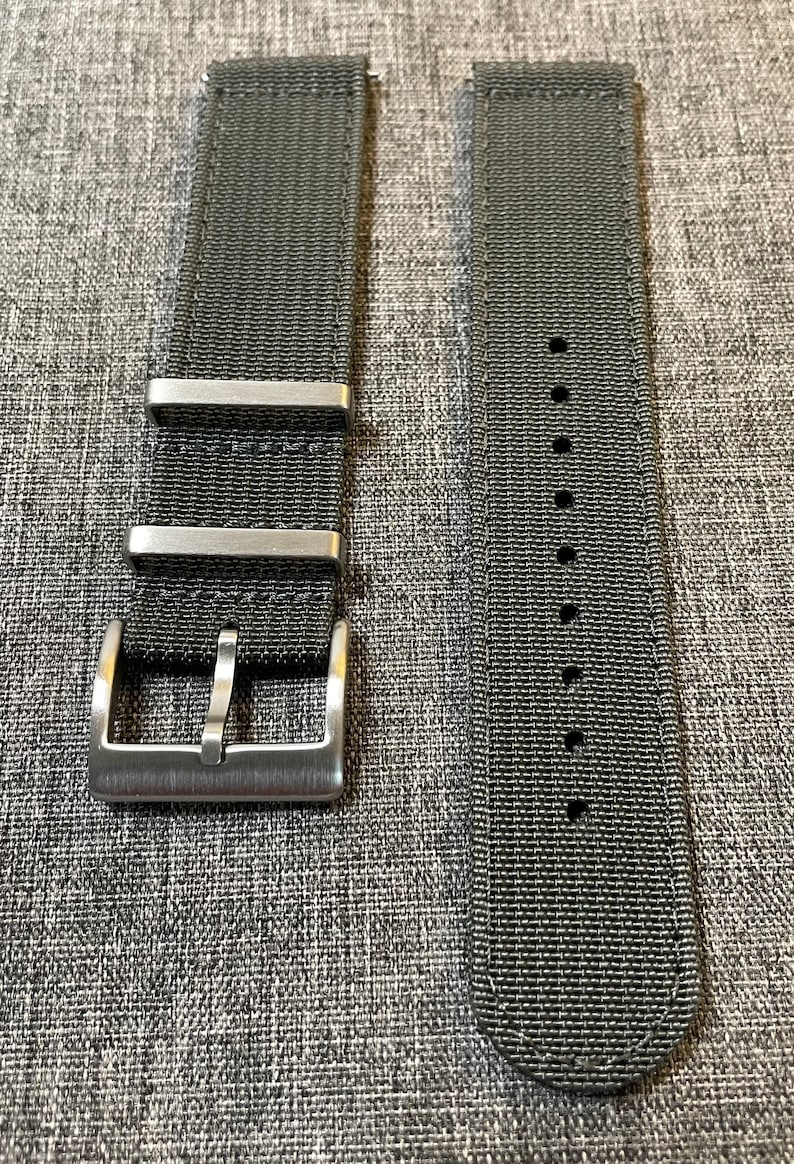 K&ouml;nnte beinhalten: Zwei schwarze Nylon-Uhrenarmb&auml;nder mit silbernen Schlie&szlig;en. Die Armb&auml;nder sind flach und haben eine gewebte Textur. Die Schlie&szlig;en haben eine geb&uuml;rstete Oberfl&auml;che.