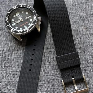 以下が含まれることがあります： 黒い腕時計と黒いラバーベルト。時計の文字盤は黒で、白いマーキングとシルバーのベゼルが付いています。ストラップはテクスチャ加工された表面とシルバーのバックルが付いています。時計には「Diver's 200m」と表示されています。