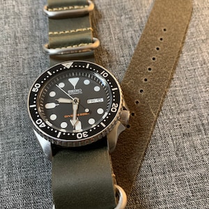 May include: A black and silver Seiko automatic dive watch with a green leather strap. The watch face has white markings and a date window that reads "FRI 5".
