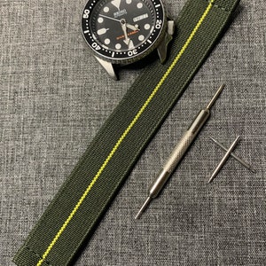 Puede incluir: Una correa de reloj verde y amarilla con una sola raya amarilla en el centro. La correa está sobre una superficie de tela gris con un reloj y herramientas de reparación de relojes.